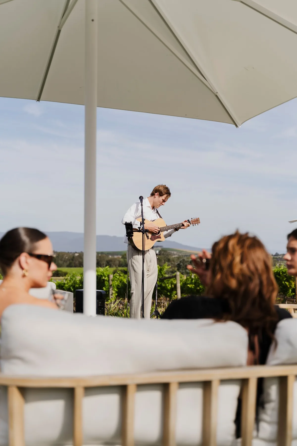 Jem Pryse playing acoustic guitar at a vineyard while guests mingle